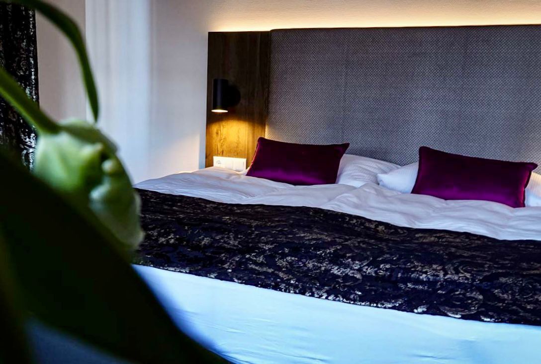 A neatly made bed with white linens and two purple pillows, a dark patterned throw, and a wall-mounted light fixture.