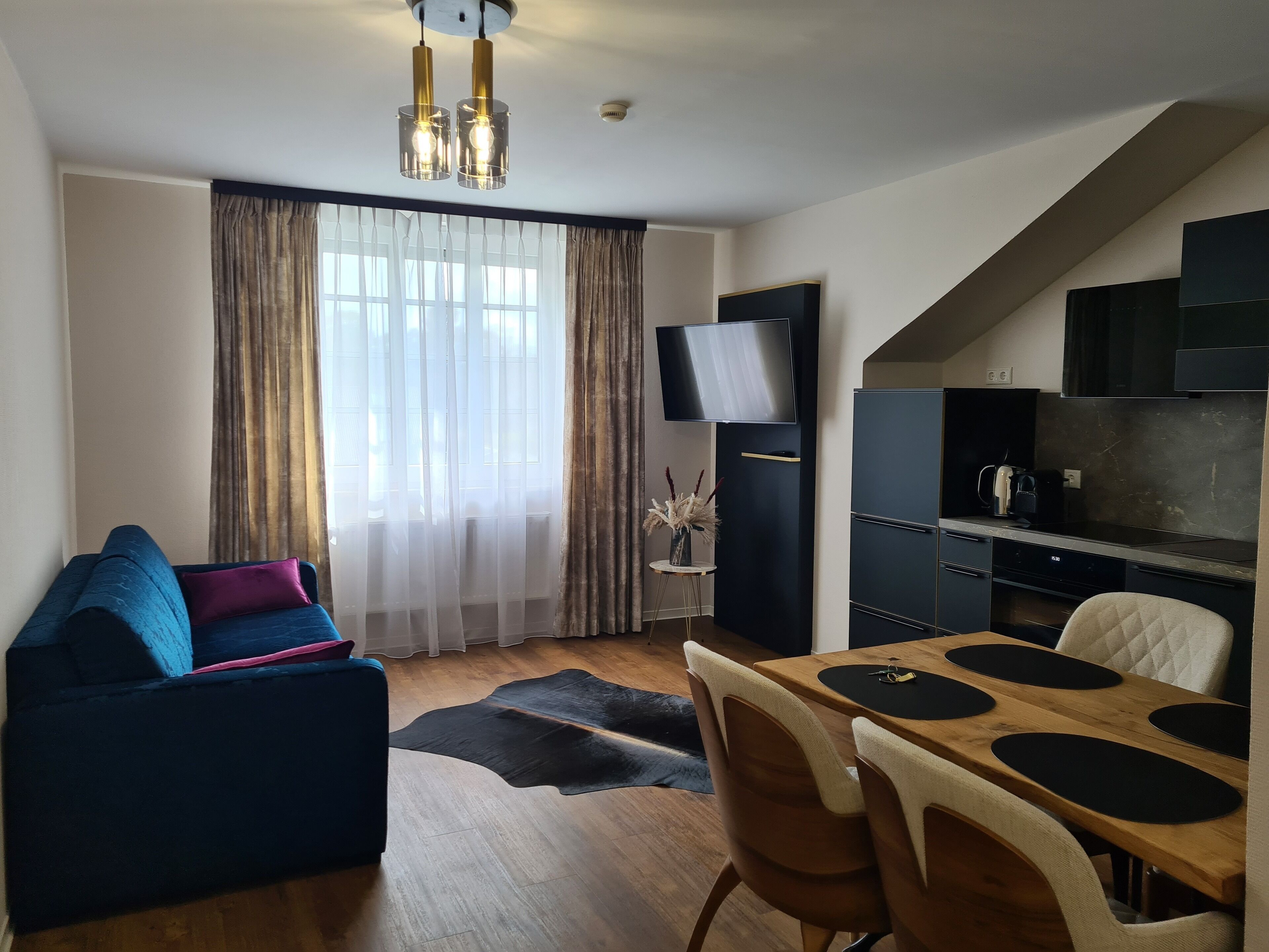 A modern living room with a sofa, a dining table with chairs, a TV, and a kitchen area with cabinets and appliances.