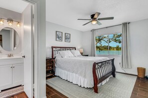 Un dormitorio con cama, ventilador de techo, ventana con vistas, mesita de noche y otro velador.
