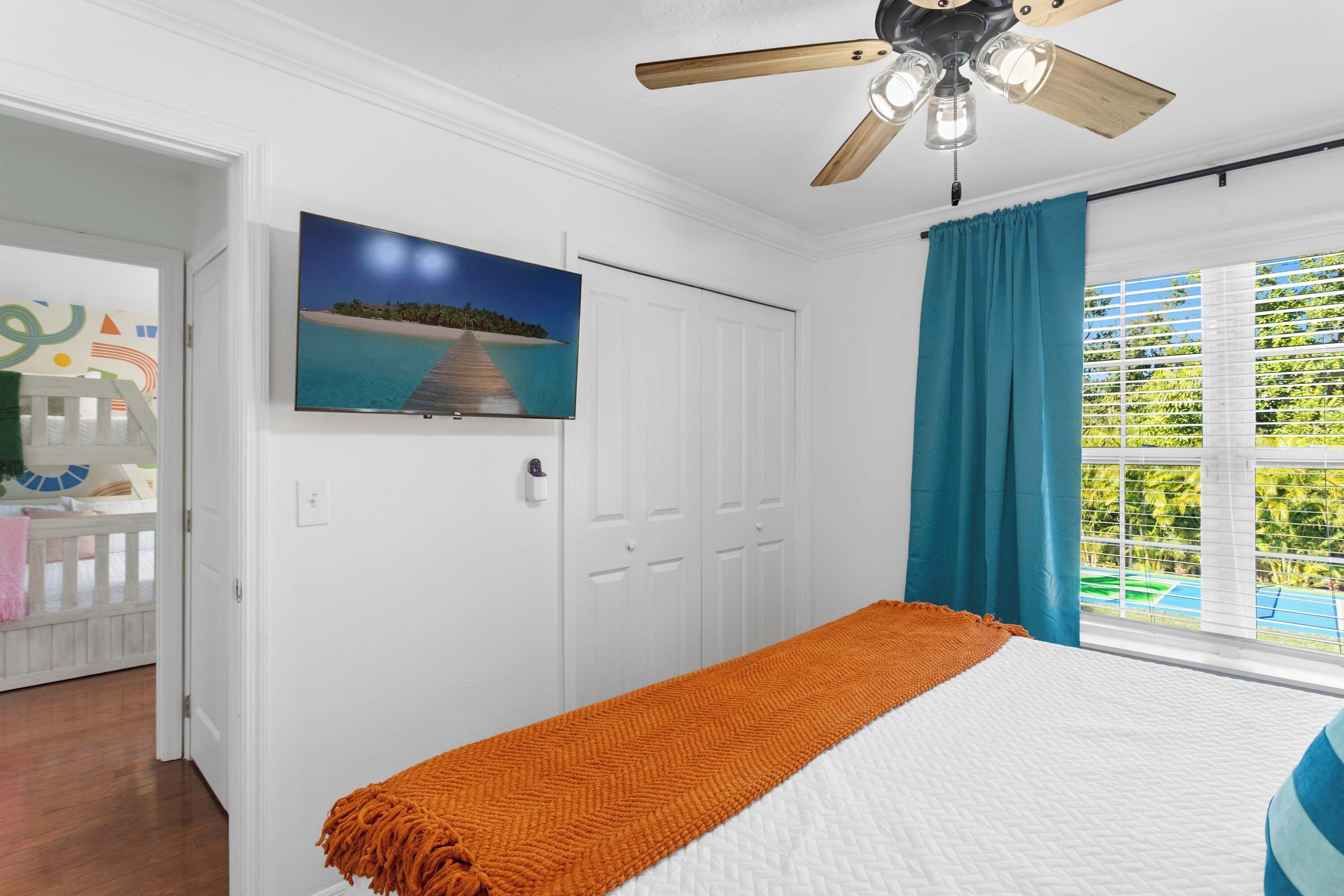 Un dormitorio con cama, ventilador de techo, televisor en la pared y ventana con cortinas.