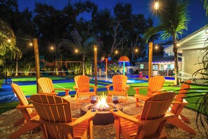 An outdoor seating area with wooden chairs around a fire pit, a pool, and a children's play area in the background.