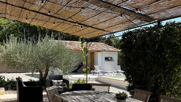 An outdoor patio with a wooden table and chairs, a pergola overhead, and a small building in the background. - Provencal house with heated pool and garden - sleeps 6 (Saint-Didier)