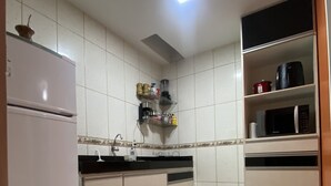 A kitchen with white tiled walls, a refrigerator, a sink, and shelves with kitchen appliances and utensils. - AconchegoF (Goiânia)
