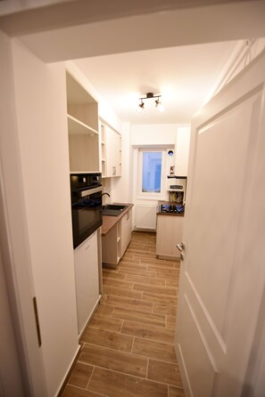 Enter a cozy space with a compact kitchen featuring white cabinetry, a black oven, and a sink, surrounded by a herringbone-patterned floor.