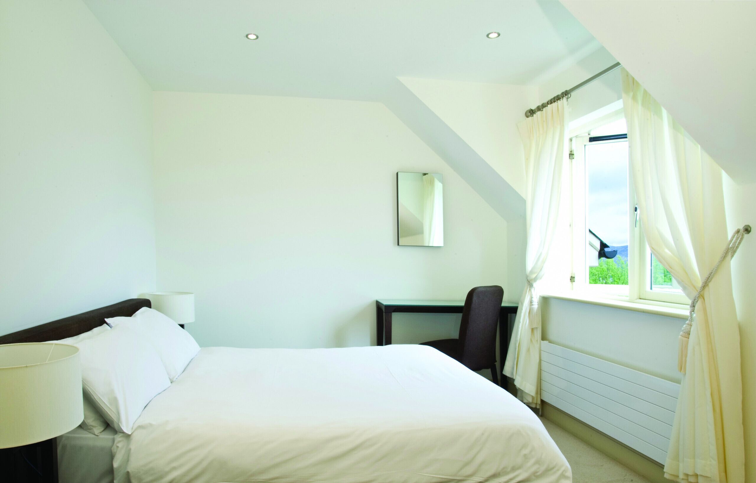 Habitación de hotel con cama, escritorio, silla, lámpara, ventana con cortinas y espejo en la pared.