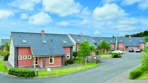 A row of red houses with grey roofs, a paved driveway, and a grassy area with a sign. - Ring of Kerry Holiday Cottages (Greenane)