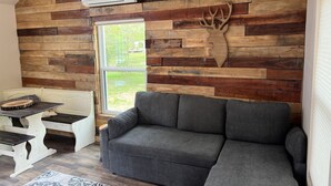 Ein gemütliches Wohnzimmer mit grauem Sofa, Holzvertäfelung, einer Dekoration mit Hirschkopf und einem Fenster mit Blick auf einen Garten.