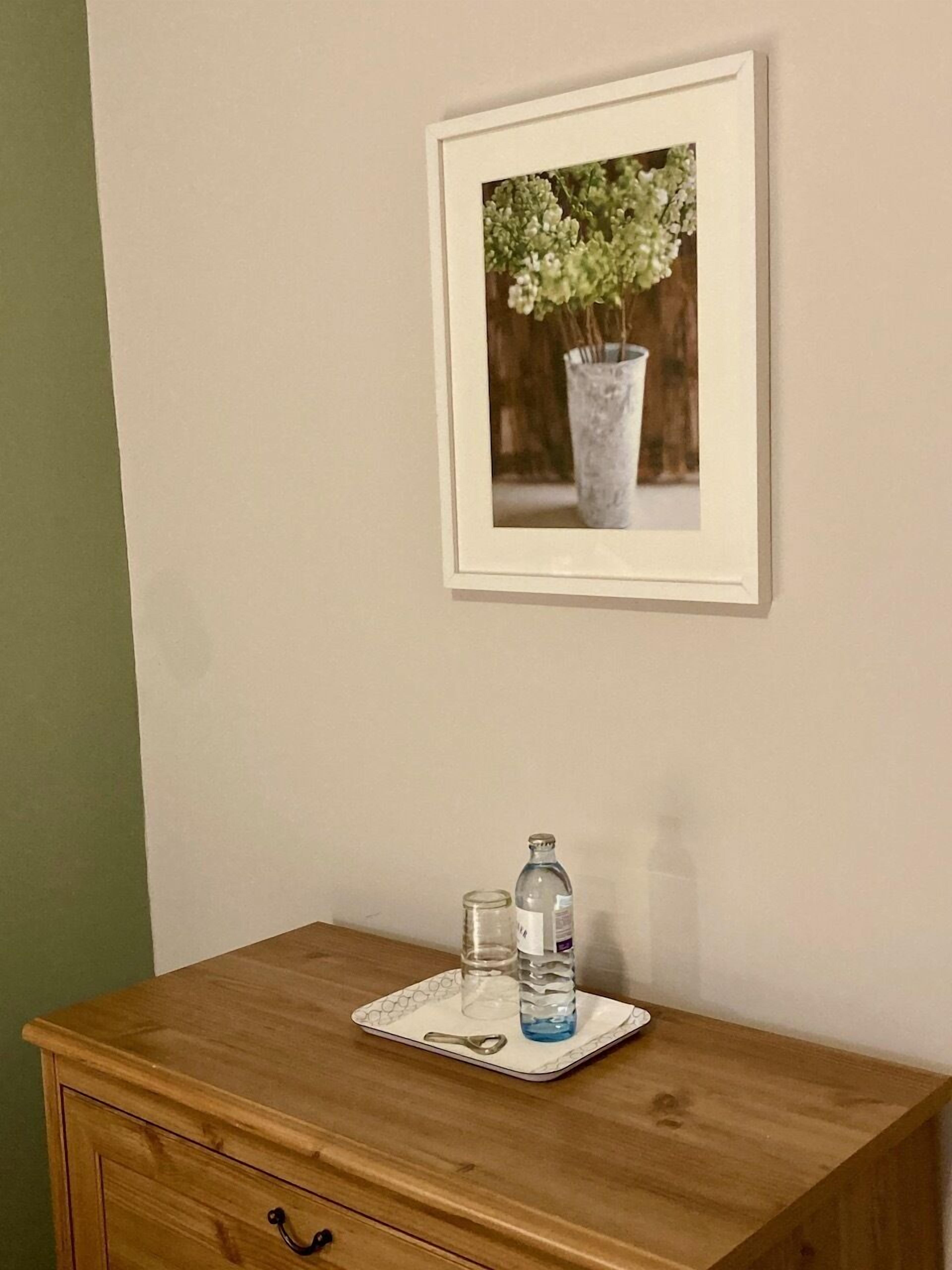 Eine Holzkommode mit einer Ablage, auf der sich eine Wasserflasche und ein Glas befinden. Darüber hängt ein gerahmtes Bild mit Blumen an der Wand.