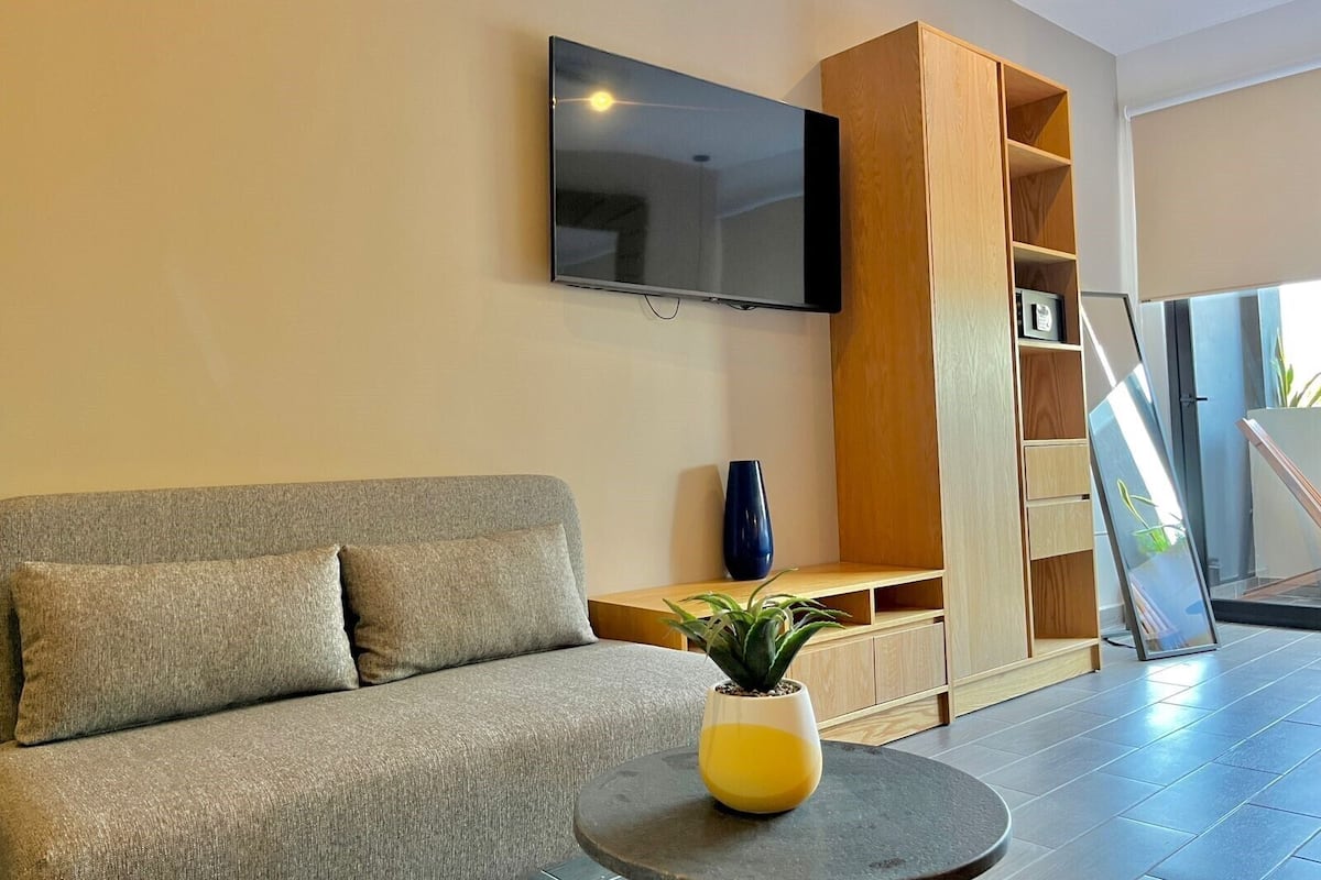 Een moderne woonkamer met een grijze bank, een wandgemonteerde televisie, een houten kast en een potplant op een ronde tafel.
