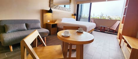 Habitación de hotel con cama, sofá, una silla de madera, una mesita con una taza y un balcón con vistas.