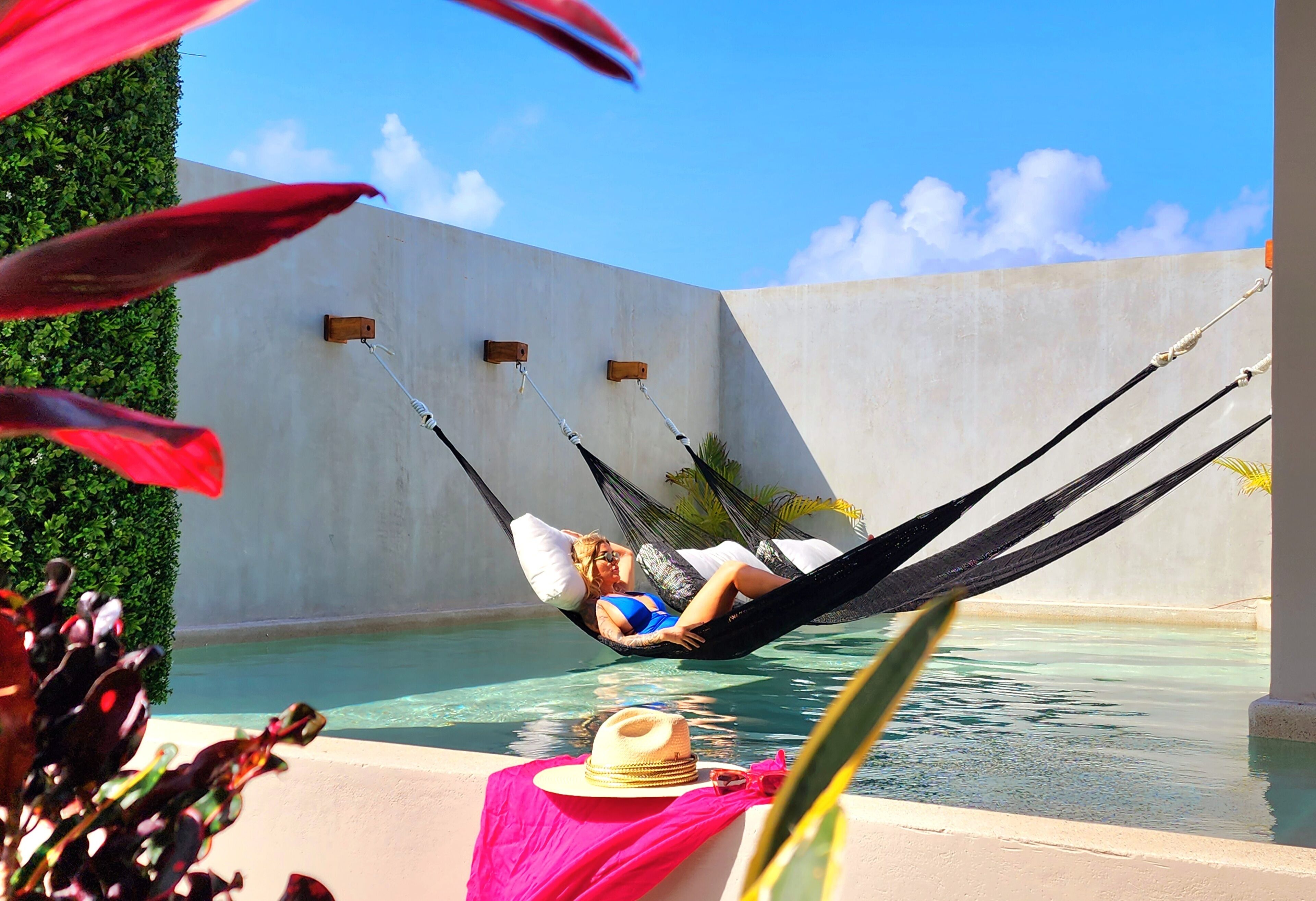 A person relaxing in a hammock by a pool with a hat and sunglasses on the side.