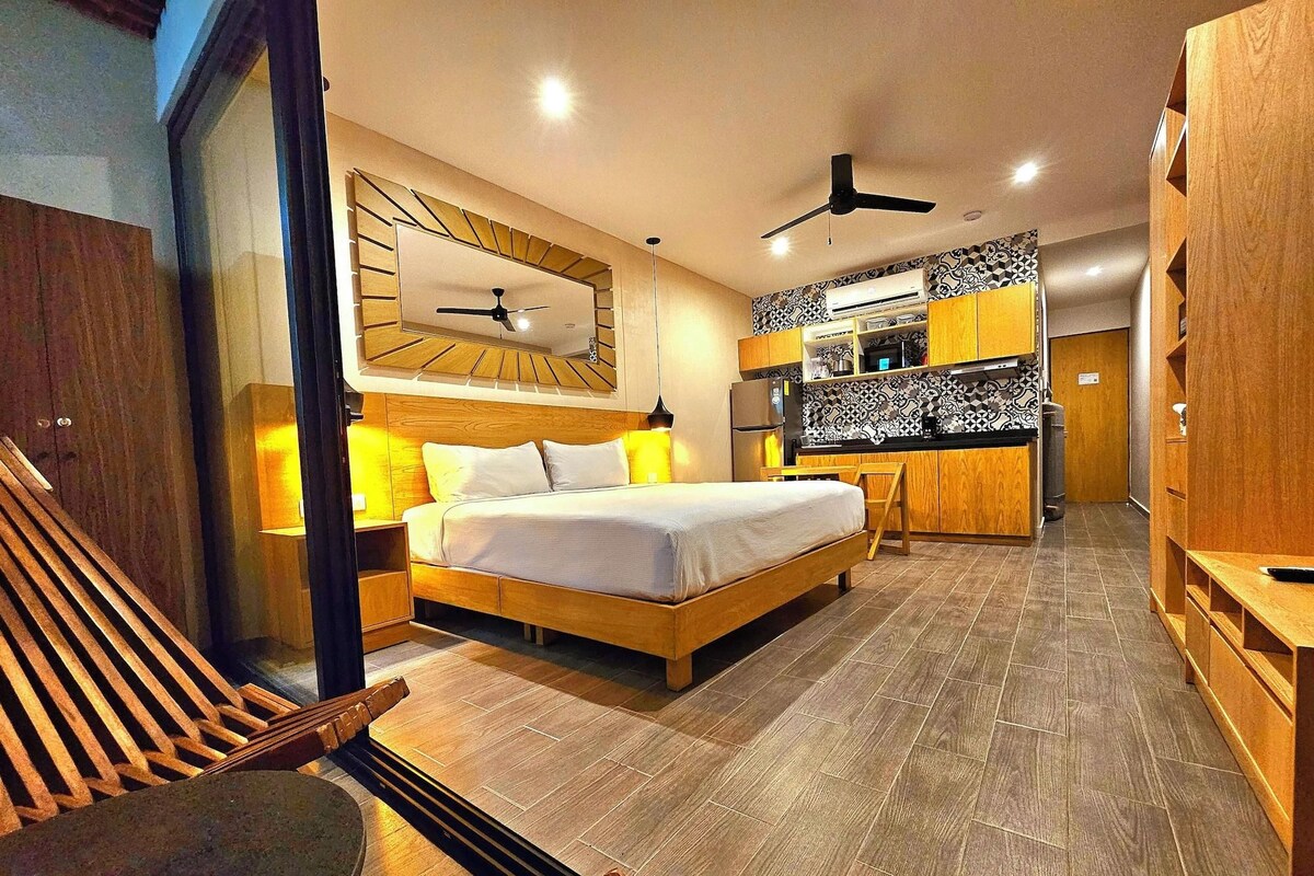 Een moderne hotelkamer met een bed, nachtkastje, lamp en een geïntegreerd keukenhoekje met een magnetron, koelkast en kastjes.