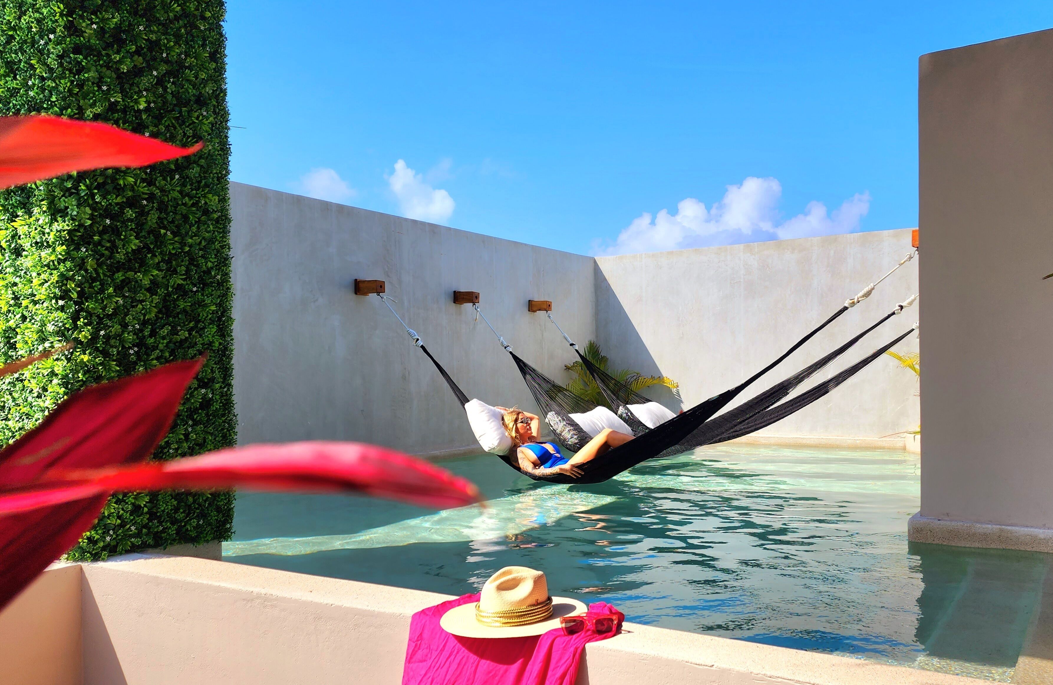 A person relaxing in a hammock by a pool with a hat and sunglasses on a towel.