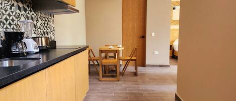 A kitchen with a black countertop, wooden cabinets, a coffee maker, and a blender. There is a wooden dining table with two chairs. An air conditioner is mounted on the wall, and a wooden door is visible in the background.