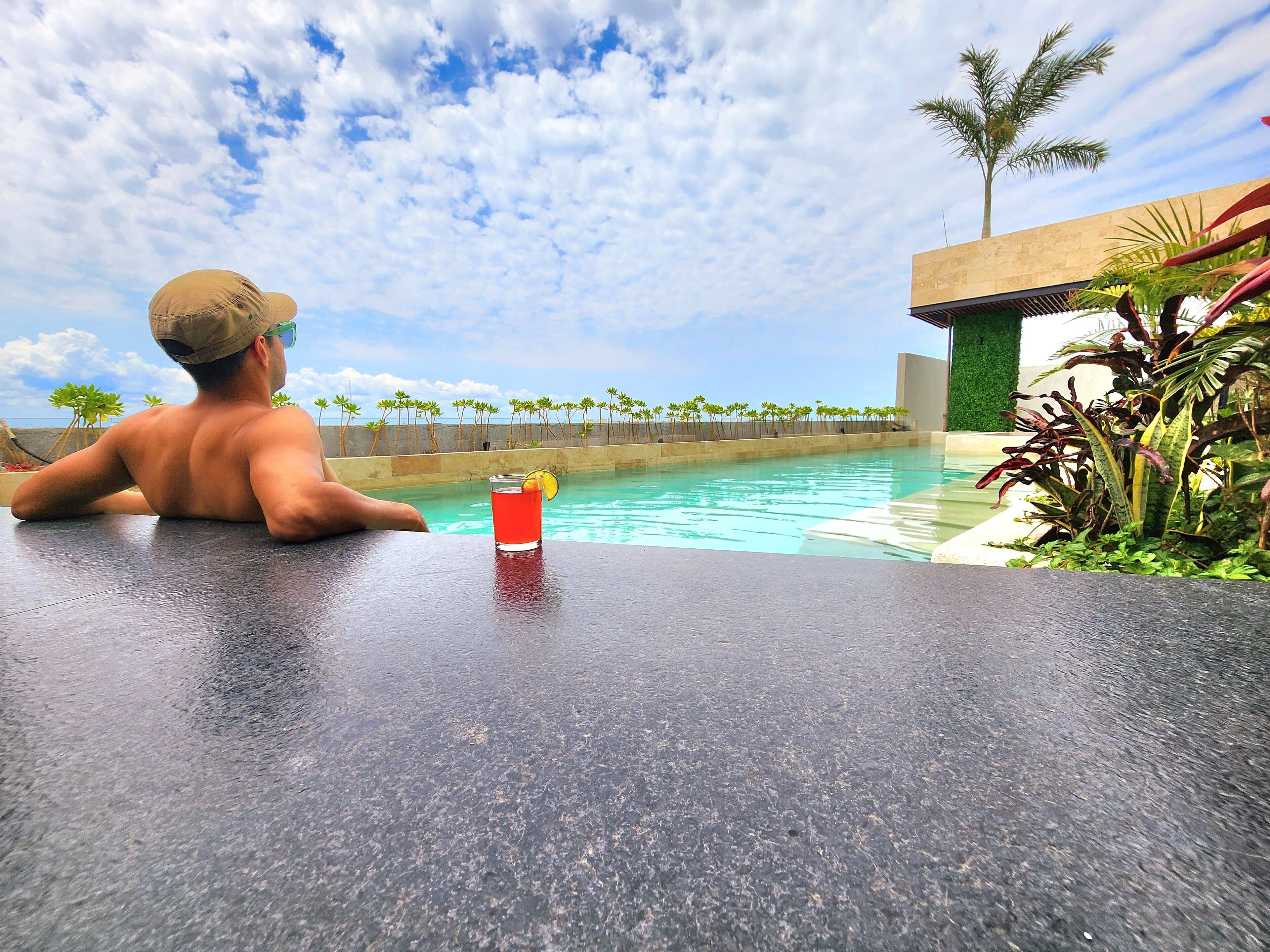 A man relaxing by a pool with a drink on the table, overlooking a pool area with palm trees and a clear sky.