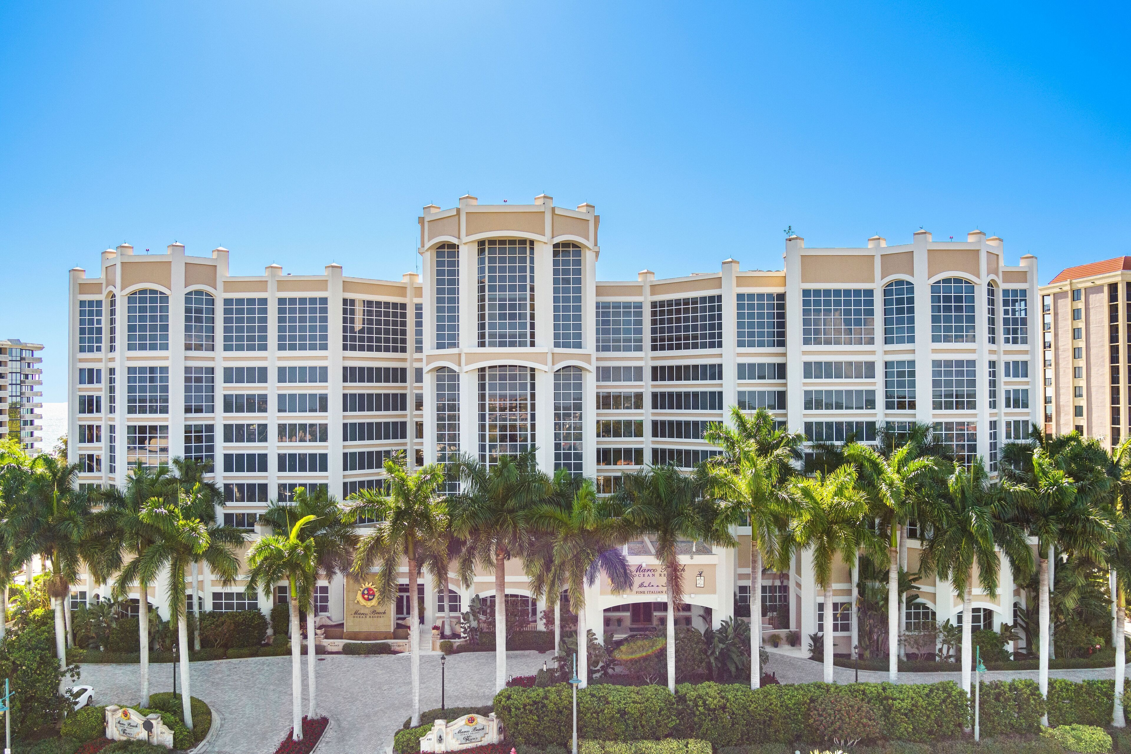 A multi-story hotel with a symmetrical facade, large windows, and palm trees in the foreground.