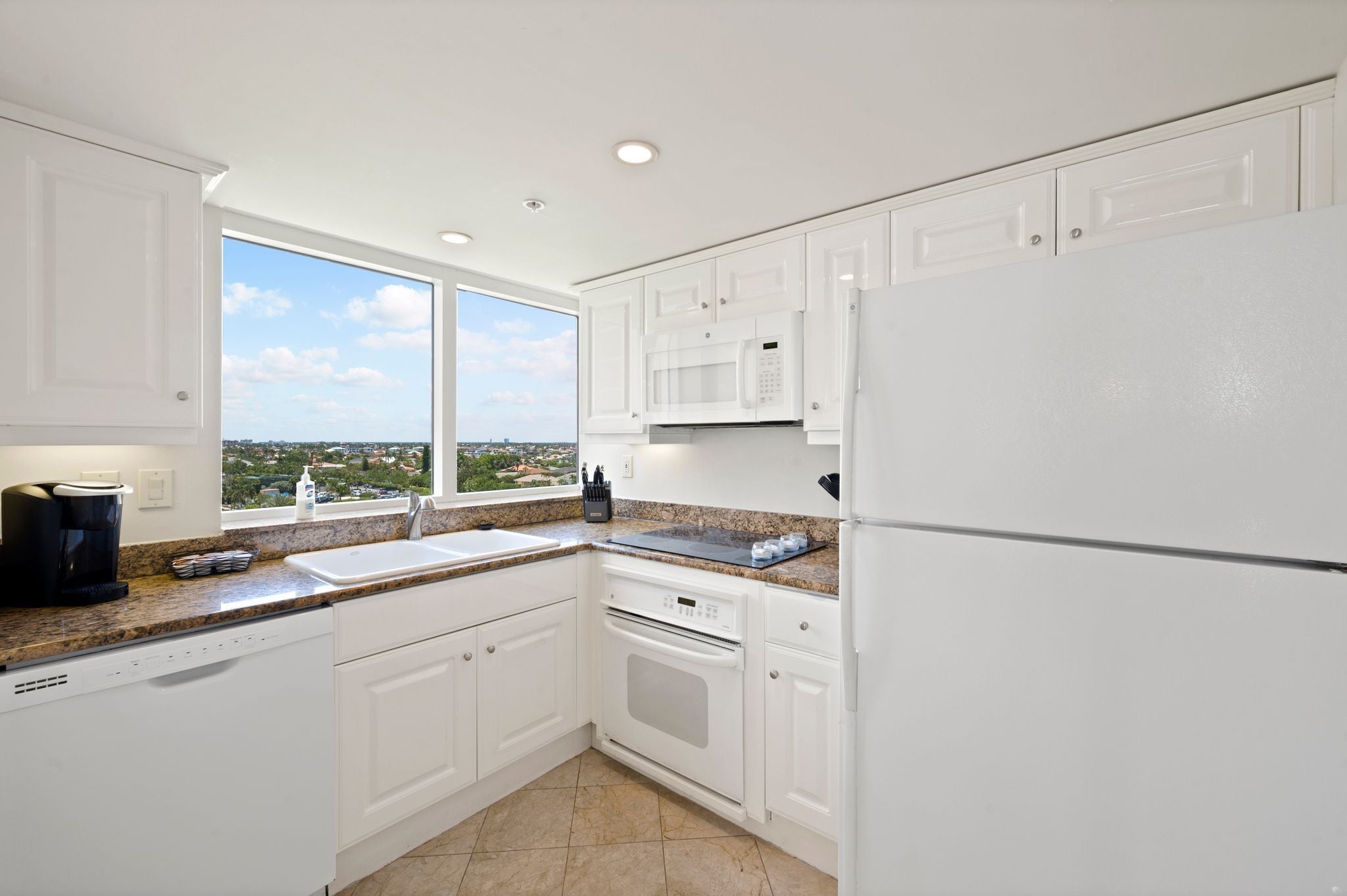 A modern kitchen with white cabinets, a built-in microwave, a stainless steel refrigerator, and a window offering a city view.