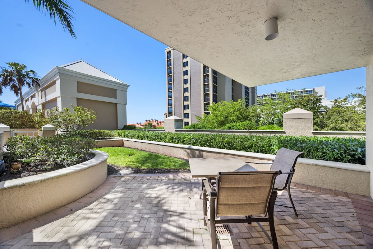 An outdoor patio with a table and chairs, surrounded by greenery and a building in the background.