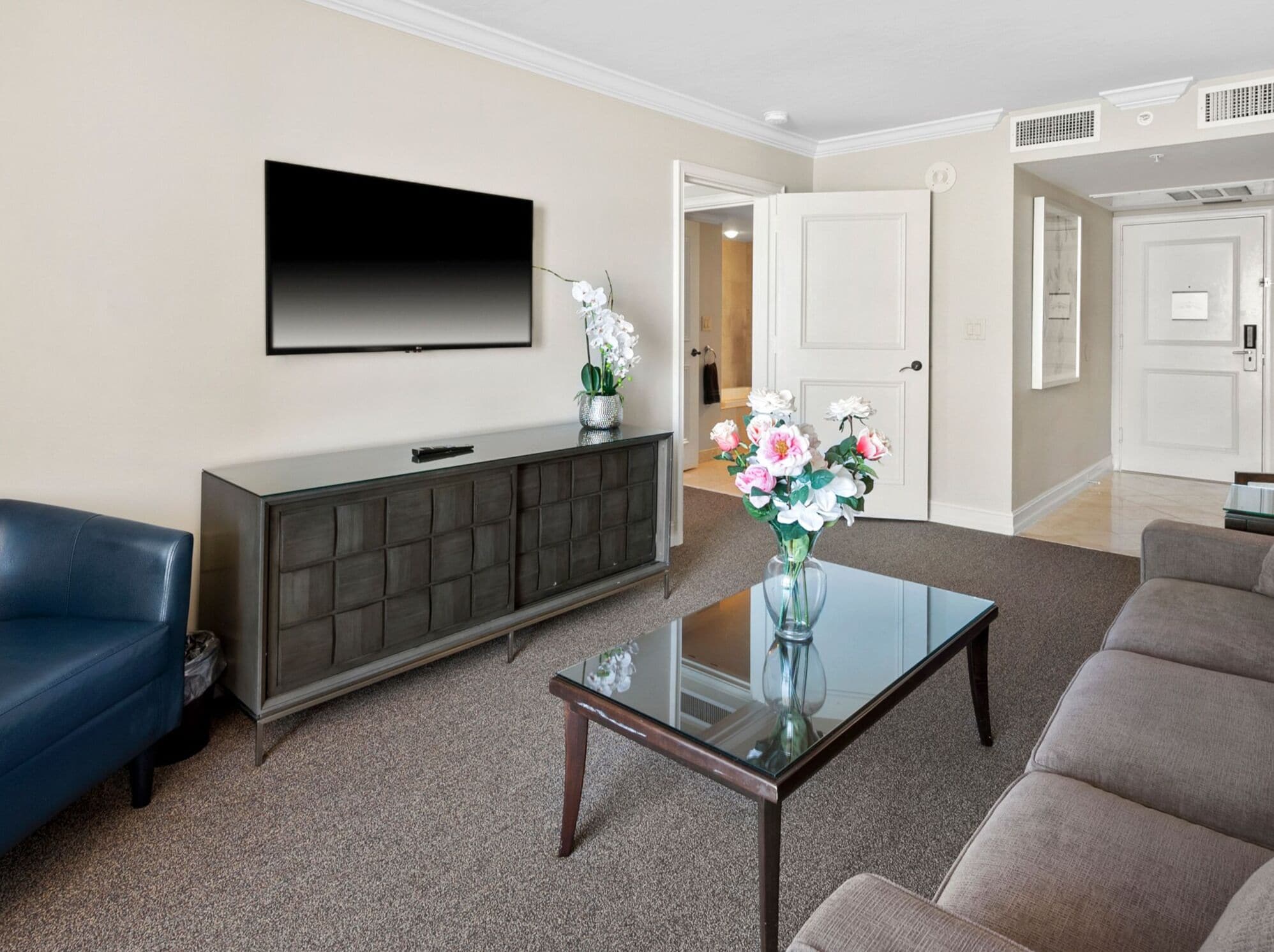A living room with a flat-screen TV mounted on the wall, a dark-toned console table, a glass coffee table, a blue sofa, and a beige sofa.