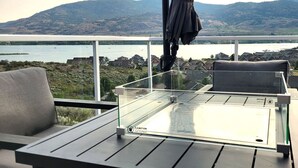 Un balcon avec une table en verre, deux chaises et une vue sur un lac et des collines.