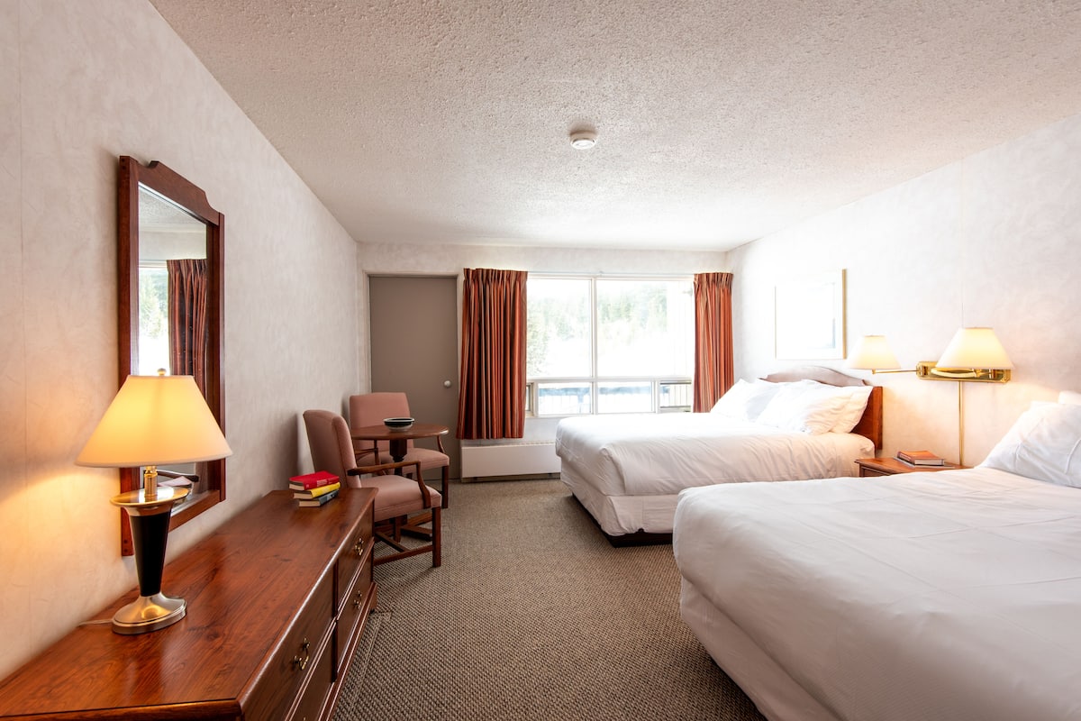 Quarto de hotel com duas camas, uma mesa com abajur, uma cadeira e vista para o exterior pela janela.