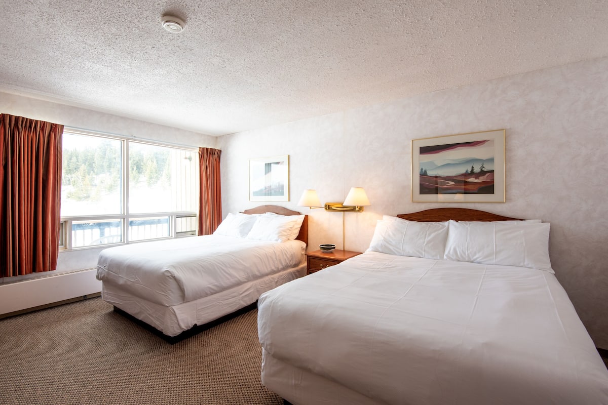 Quarto de hotel com duas camas, uma janela grande com cortinas, um quadro na parede e uma luminária na mesa de cabeceira.