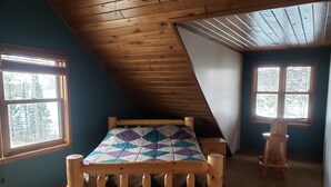 Una habitación con techo inclinado, una cama de madera con colcha estampada, dos ventanas con persianas y un pequeño taburete de madera.