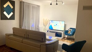 Un salón con un sofá color beige, un sillón azul, un televisor sobre un mueble blanco y una ventana con cortinas translúcidas.