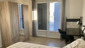 Un dormitorio con cama, escritorio, silla, espejo y vistas a través de una ventana con cortinas.
