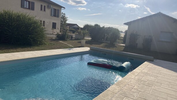 Una piscina con un objeto flotando, rodeada de un solárium empedrado y contigua a un edificio.