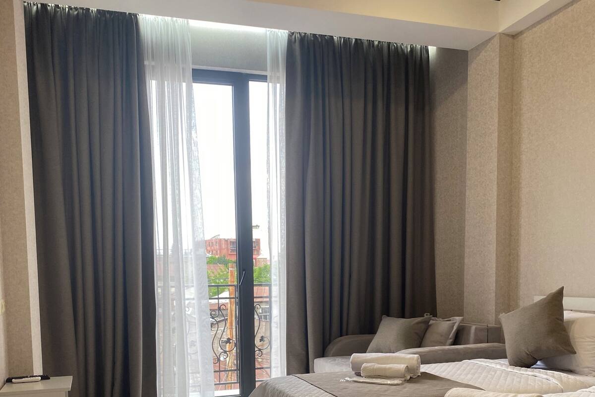 A modern hotel room with a large bed, a bedside table, and a view through a glass door to the outside.