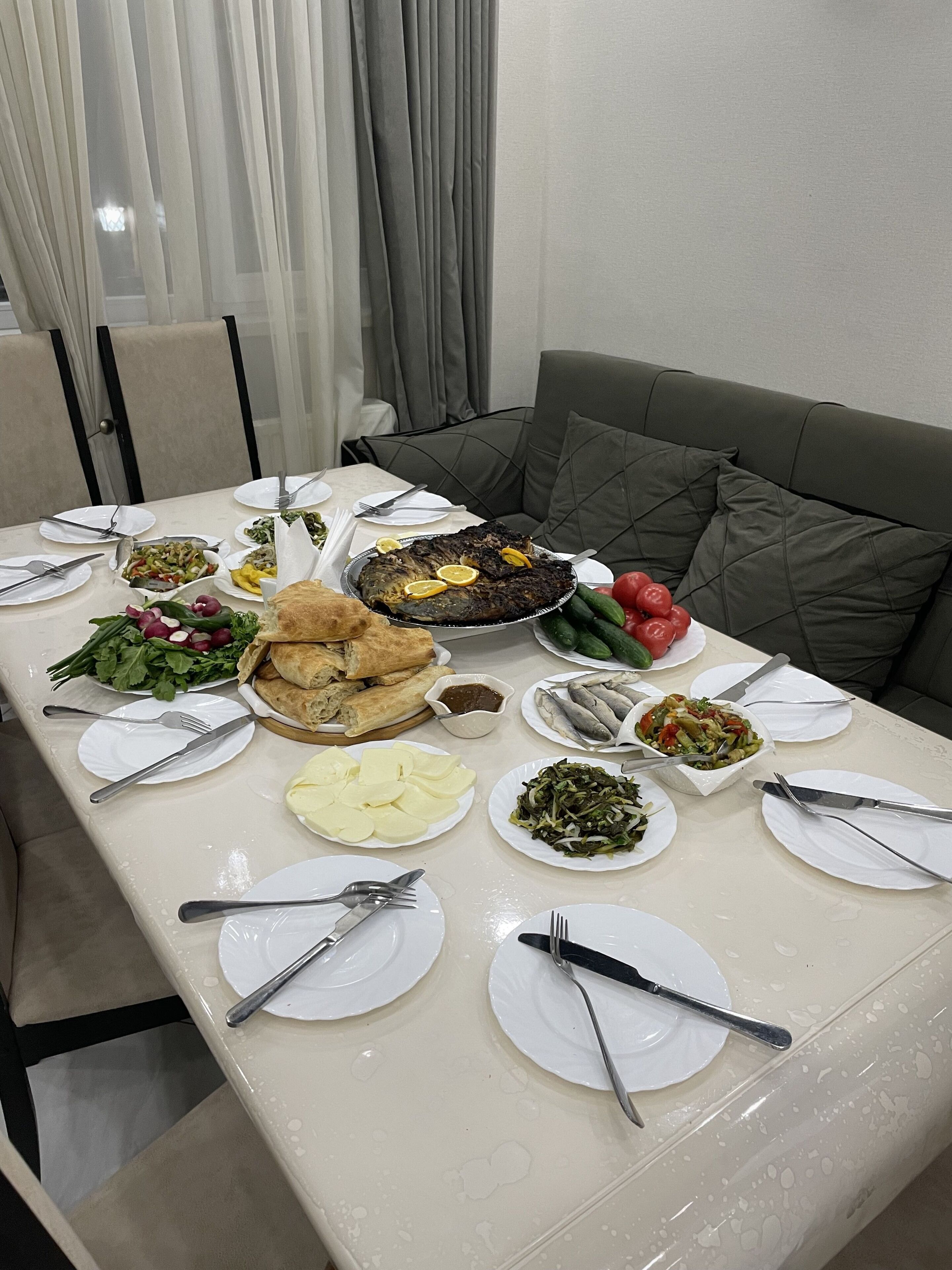 Une table à manger dressée avec divers plats, notamment du poisson, des légumes et du pain, dans un espace repas moderne agrémenté d’un canapé gris et de rideaux.
