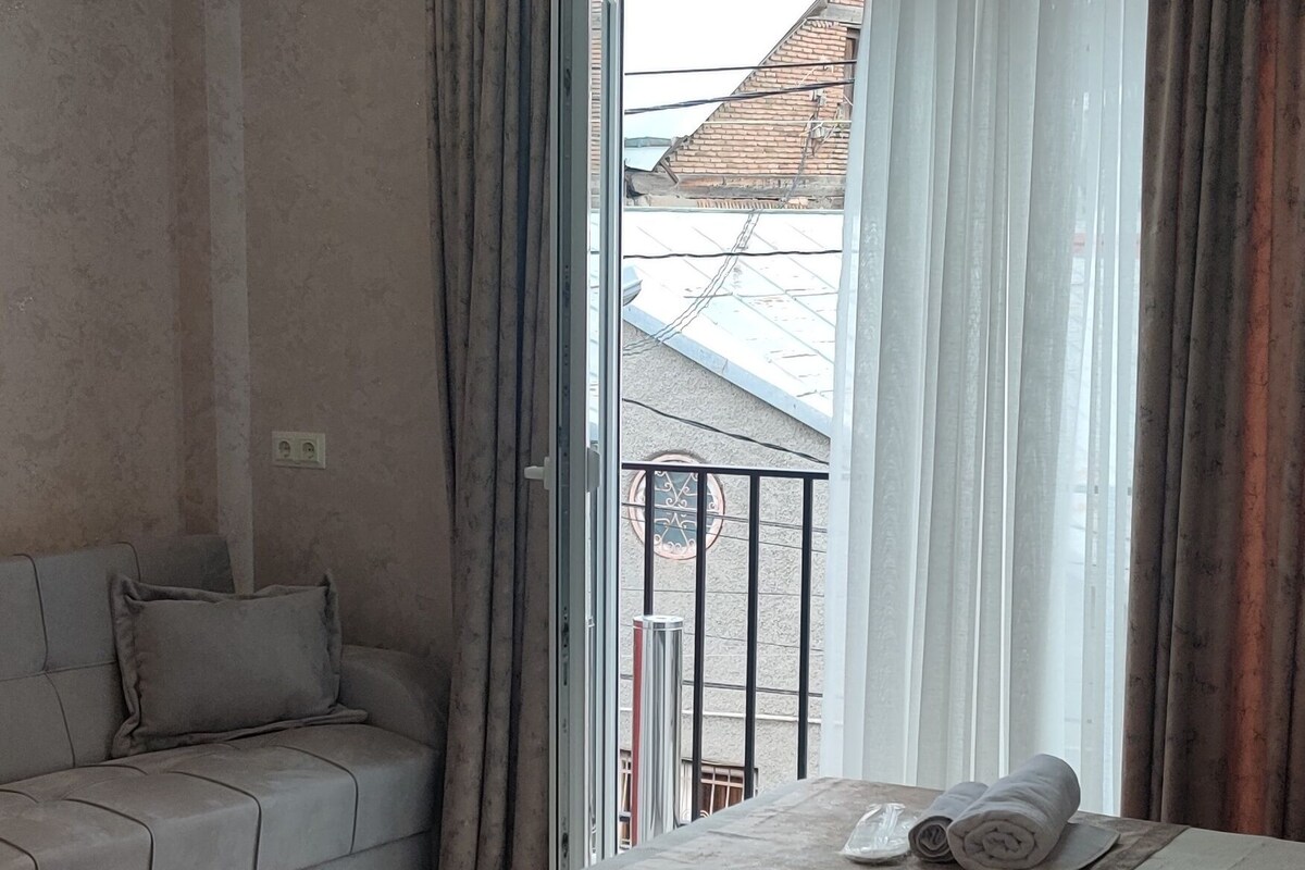 A hotel room with a sofa, a bed, a table, and a chair. There is a window with curtains, and a balcony is visible outside.