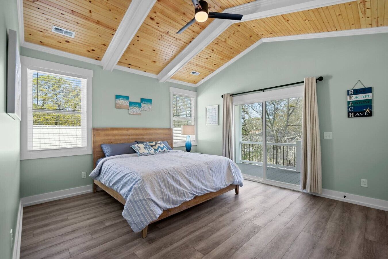 Een slaapkamer met een houten plafond, een bed met een blauw-witte spreidekker en een groot raam met uitzicht op bomen.