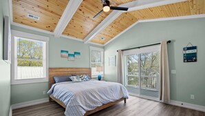 Una camera da letto con soffitto in legno, un letto con copriletto blu e bianco e un'ampia finestra vista alberi.