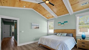 Una camera da letto con soffitto in legno, un letto con copriletto a righe, un comodino con una lampada e una finestra con persiane.