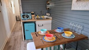 Una cocina compacta con un refrigerador azul, una mesa de comedor de madera con tazones y platos coloridos, y un lavamanos con diversos objetos.
