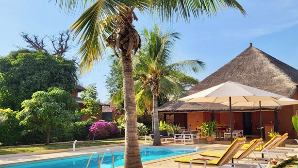 A poolside area with lounge chairs, a thatched-roof building, and a palm tree. - Villa Sarrène (Nianing)