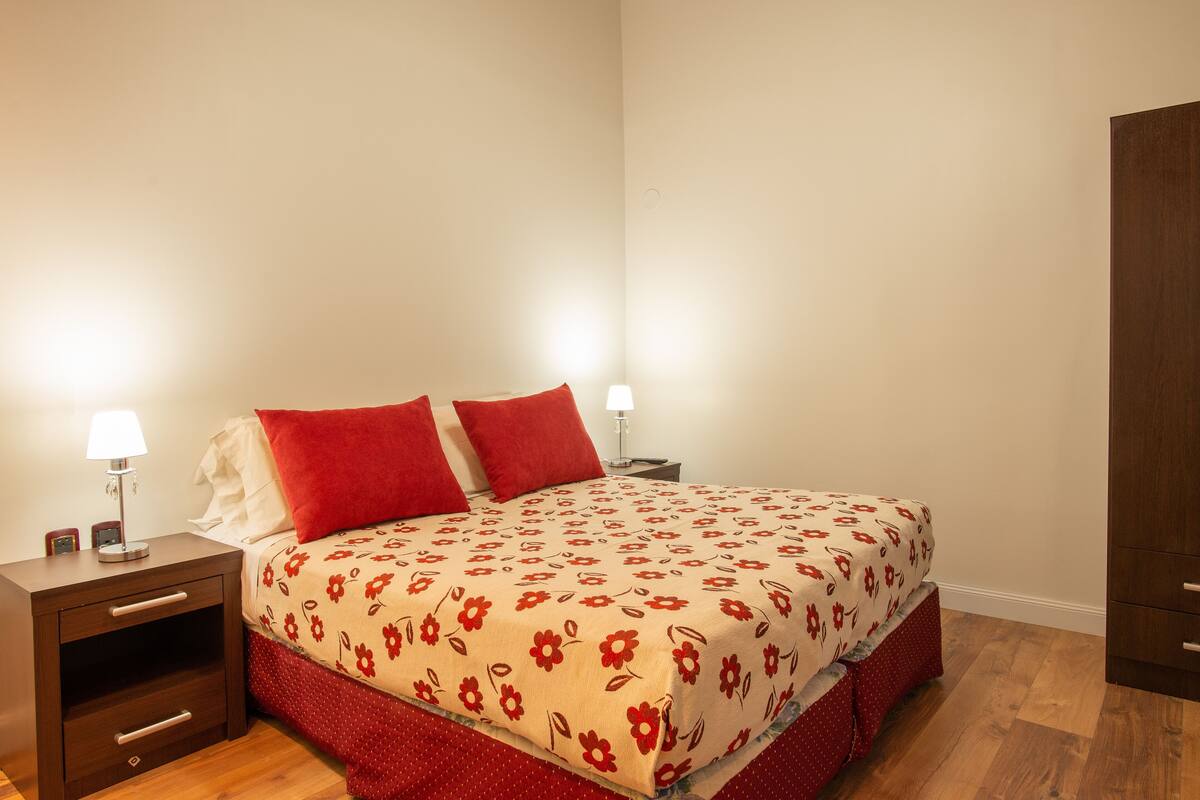Un lit bien fait, recouvert d’une couvre-lit à motifs floraux, avec deux oreillers rouges et deux lampes de chevet posées sur une table de nuit en bois.