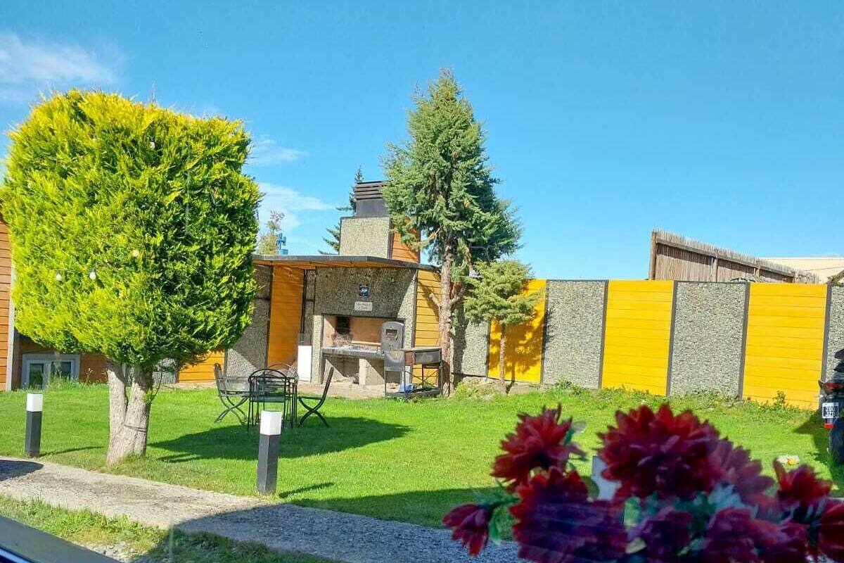 On voit un bâtiment avec une façade jaune, un petit jardin avec un arbre, et un pot en métal rempli de fleurs au premier plan.