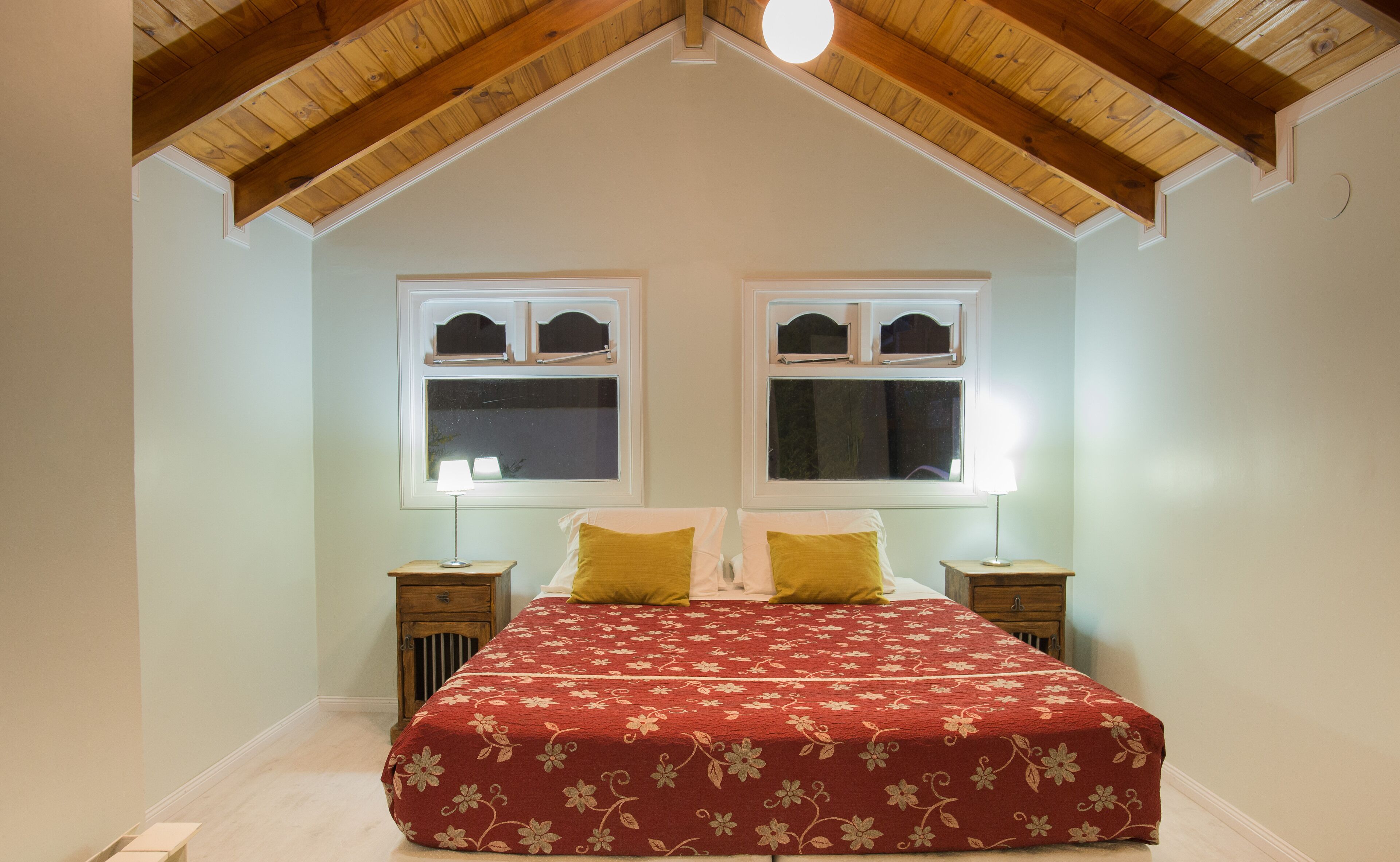 A bedroom with a sloped ceiling, two windows with shutters, a bed with a floral bedspread, and bedside lamps.