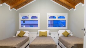 Ein Zimmer mit zwei Betten, jeweils mit einem weißen Nachttisch und einem Fenster. Das Zimmer verfügt über eine schräge Holzbalkendecke und helle Wände.