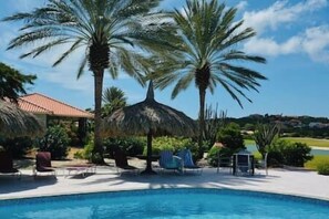 A swimming pool with lounge chairs and palm trees, surrounded by a building with a red roof. - VIP Blue Bay Beach Villa 33 (Sint Michiel)