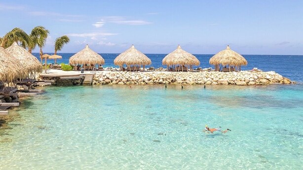 Una playa con agua turquesa cristalina, construcciones de techo de paja y un muelle rocoso.