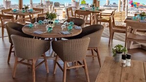 Un restaurante frente a la playa con mesas redondas, sillas de mimbre y plantas en macetas.