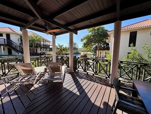 A covered outdoor area with wooden flooring, lounge chairs, and a pool in the background. - VIP Blue Bay Beach Villa 33 (Sint Michiel)