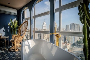A room with a large window, a bathtub, a wicker chair, and a cactus plant. - Vossa Bossa Sky Mirante (São Paulo)