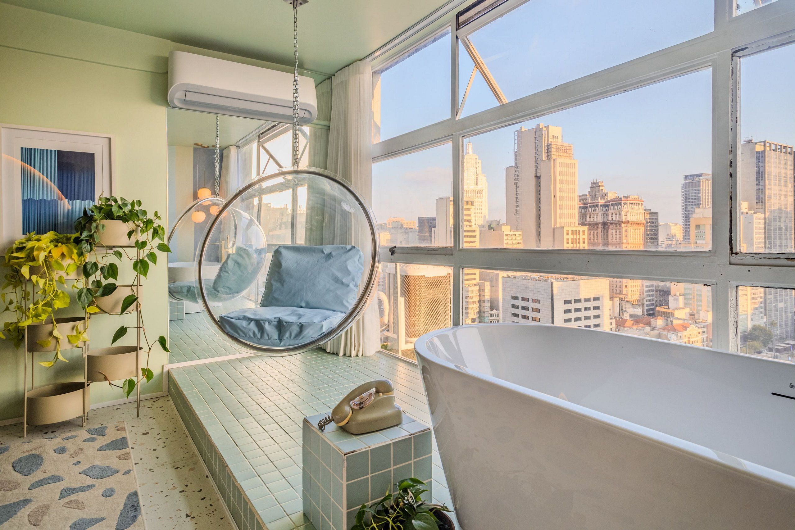 A room with a view of a cityscape, a hanging chair, a bathtub, and a telephone on a small table.