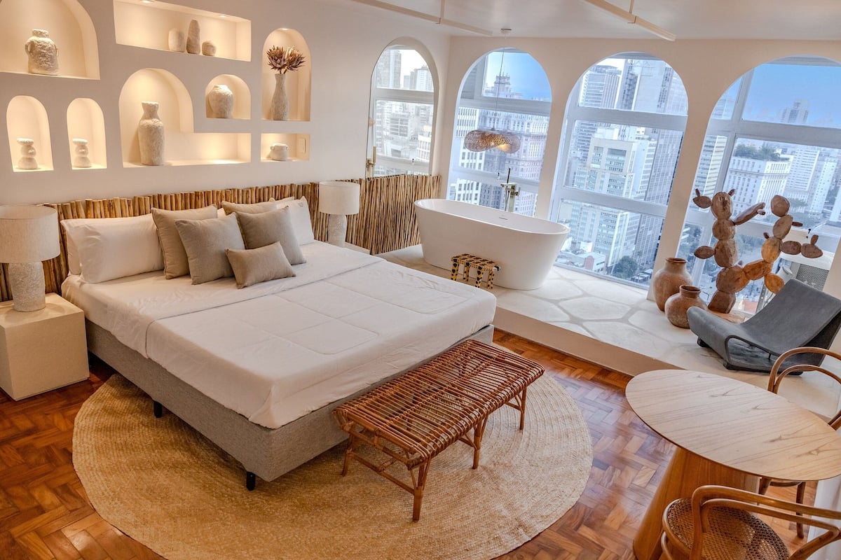 Un dormitorio moderno con una cama grande, una bañera y vistas de la ciudad a través de ventanales de arco.
