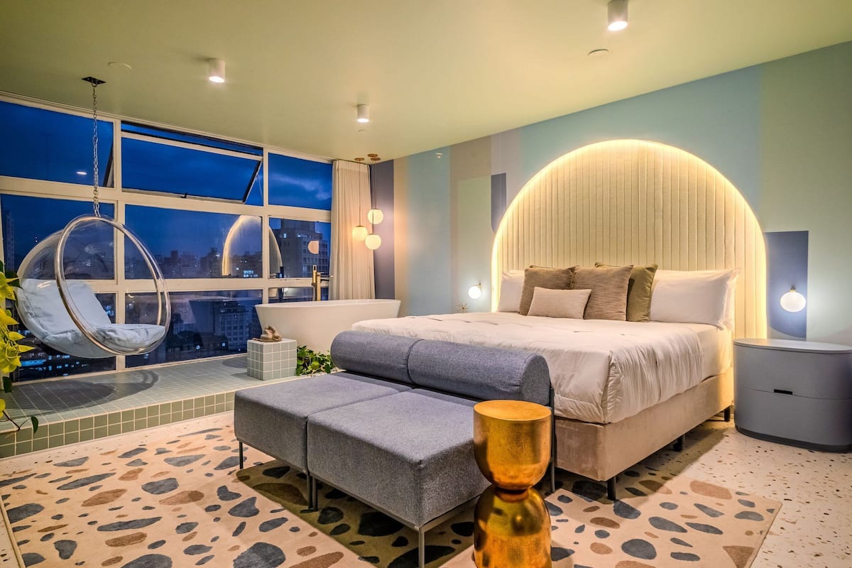 Un dormitorio moderno con una cama grande, una silla colgante, un banco y vistas de la ciudad por la noche.